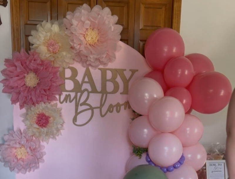 Pink arch backdrop with paper flowers, a Baby in Bloom sign, and a pink and sage balloon garland