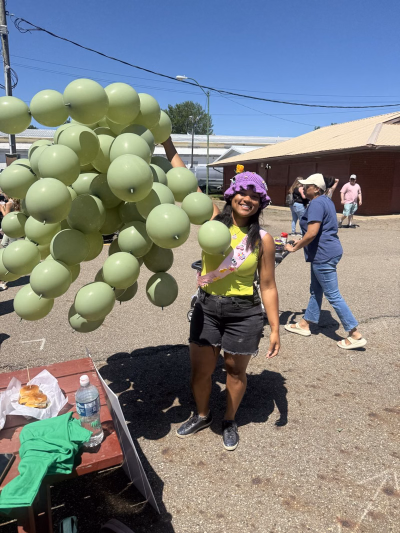 Key smiling in a purple crochet hat holding a large bunch of sage green balloons