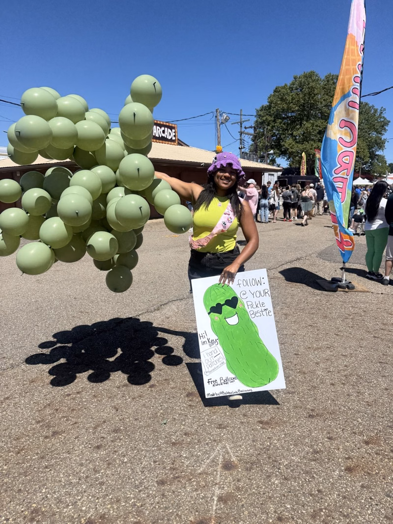 Key holding a cloud of sage green balloons at a street fair next to a painted pickle sign