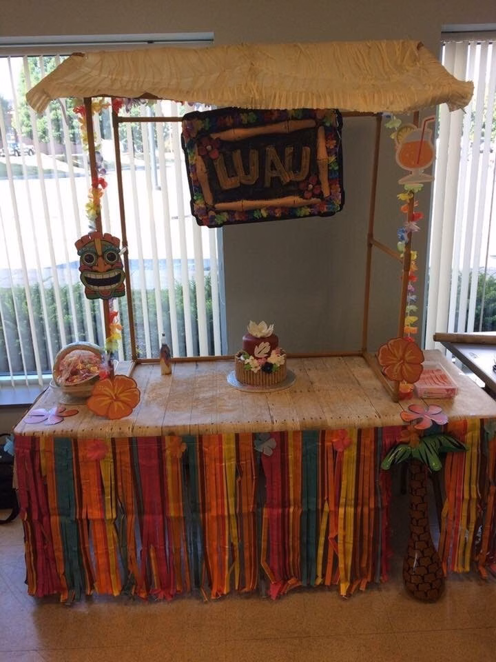 Luau themed dessert table with a bamboo style sign, tiki decor, and a colorful fringe table skirt