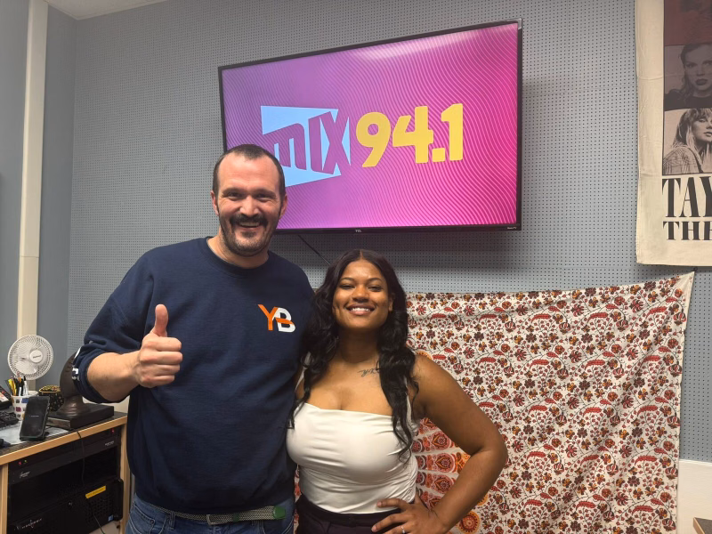 Two people smiling in a radio studio beneath a screen showing the Mix 94.1 logo, one giving a thumbs up