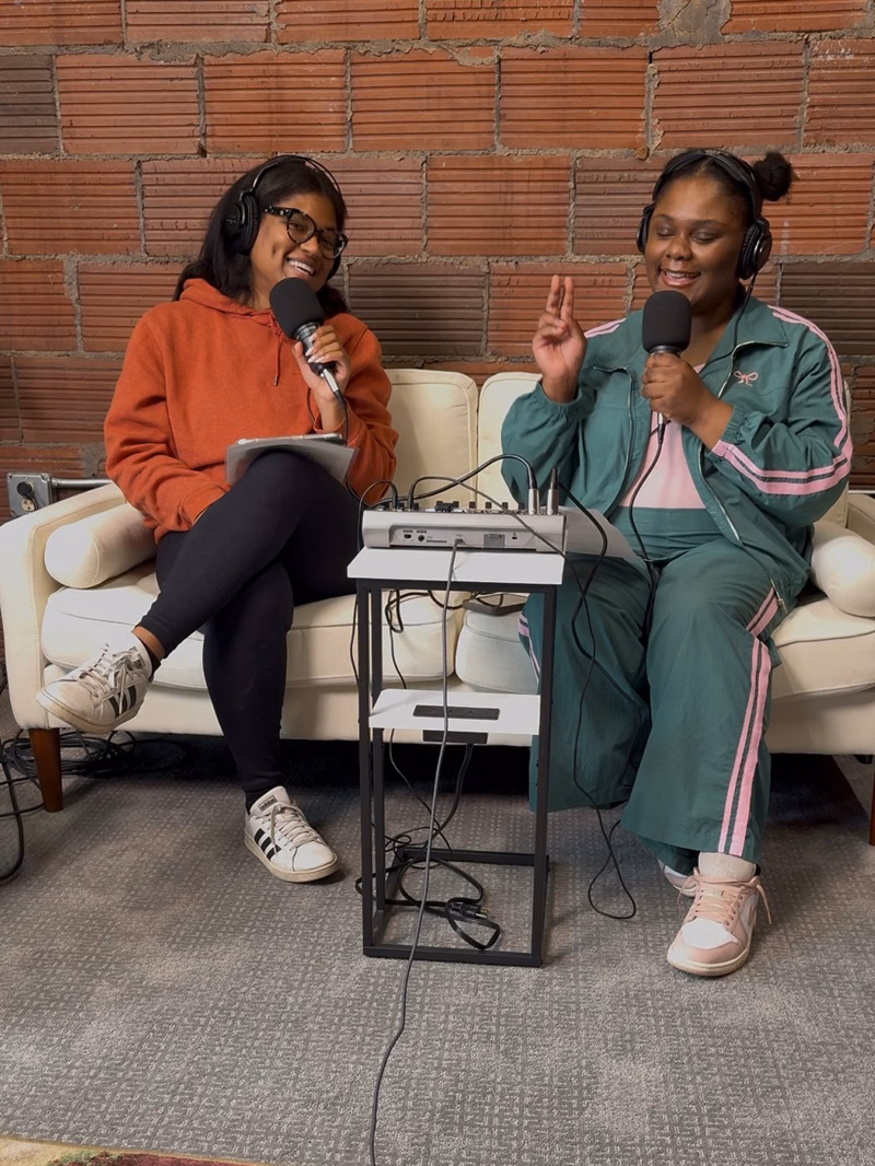 Two hosts laughing with microphones and headphones on a cream couch in front of a brick wall