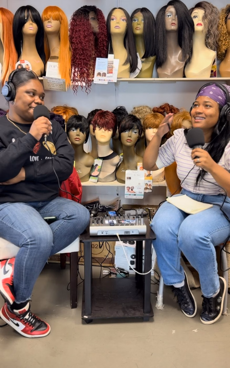 Two hosts with microphones in front of shelves of wigs in a shop