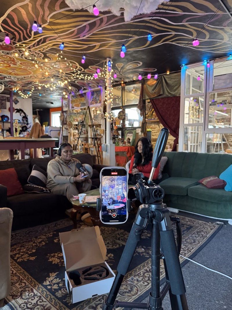 Phone on a tripod filming two hosts on couches in a colorful art studio with string lights